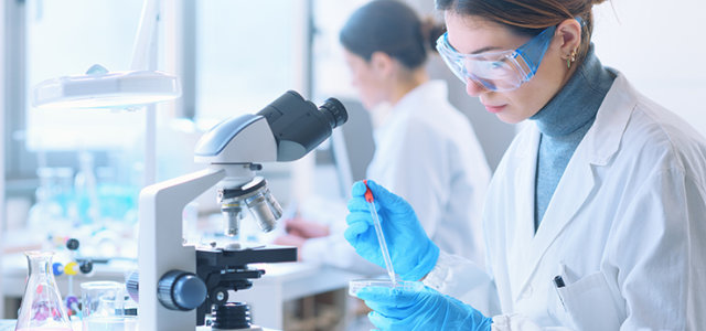 Eine Laborantin mit weißem Kittel und Schutzbrille befüllt eine Petrischale mit einer Pipette und sitzt dabei vor einem Mikroskop. (Bild:Adobe Stock)