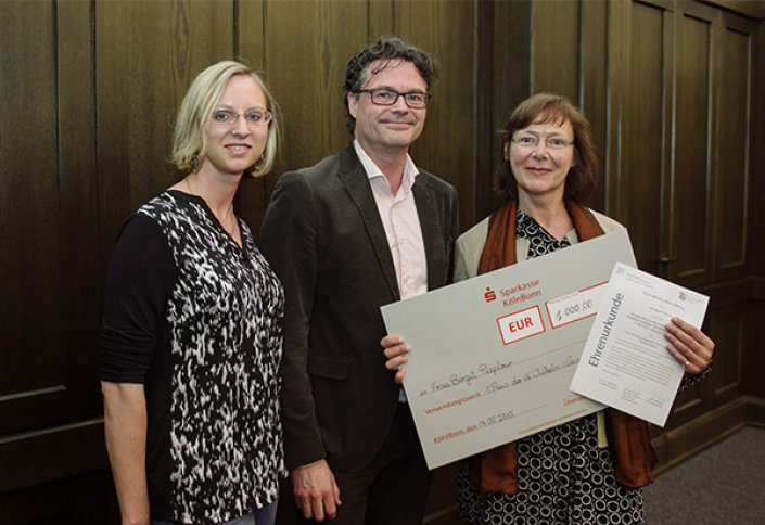 von links: Natascha Klein, Prof. Dr. Rolf Schwartmann, Preisträgerin Birgit Pieplow hält einen übergroßen Scheck und eine Ehrenurkunde in der Hand